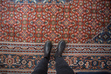 Rust field with Herati design, denim blue border details, and black boots on vintage rug.