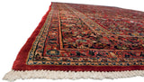 Angled view of a vintage Sarouk carpet's corner, highlighting the full red pile, ivory fringe, and floral borders.