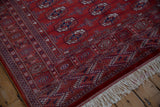 Angled corner view of a fine Bokhara rug showing its lustrous red pile, intricate borders, and ivory fringe.