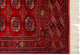 Cherry red Bokhara rug with geometric gul medallions, intricate navy border, and cream fringe.