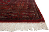 Angled view of a vintage Bokhara carpet's corner, showing the plush red pile and full ivory fringe.