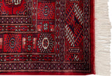Vintage Bokhara rug corner showing cherry red field, navy gul motifs, secured red selvage, ivory fringe.