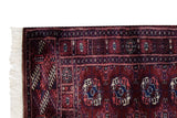 Corner of a Bokhara rug showing its full ivory fringe, intact selvage, and dense wine-red pile.