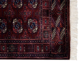 Corner detail of a fine Bokhara rug with wine red and navy blue geometric motifs next to its ivory fringe.