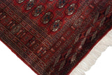 Corner of a fine Bokhara rug showing its plush red pile, side cord selvage, and ivory fringe.