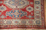 Cherry red Pakistani Caucasian rug corner with ivory border, blue geometric motifs, and white fringe.