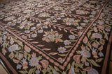 Curvilinear chainstitch floral vines and patterned border in dusty blue, mauve, and olive on dark brown.