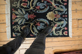 Black vintage Indian Savonnerie design rug with looped pile, vivid floral motifs, shown with person's boots.