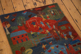 Vibrant pictorial folk art rug showing a village scene with figures, homes, and trees.