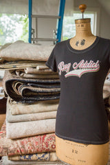 Charcoal 'Rug Addict' t-shirt on a vintage mannequin beside a stack of folded antique rugs.