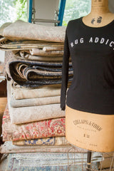 Black Rug Addict long sleeve t-shirt on a vintage mannequin beside a stack of folded vintage rugs.