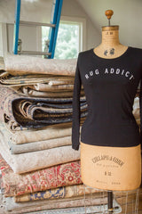 Black Rug Addict long sleeve shirt on a vintage dress form beside a tall stack of folded rugs.