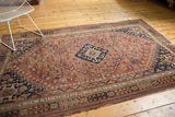 Qashqai rug corner with dusty rust field, intricate navy motifs, and visible wear.