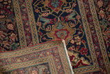 Persian Kerman rug corner revealing dense hand-knotting, white fringe, and classic navy, red floral design.
