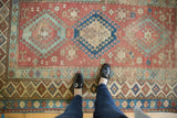 Distressed vintage Caucasian rug, showing geometric medallions, worn texture, and feet for scale.